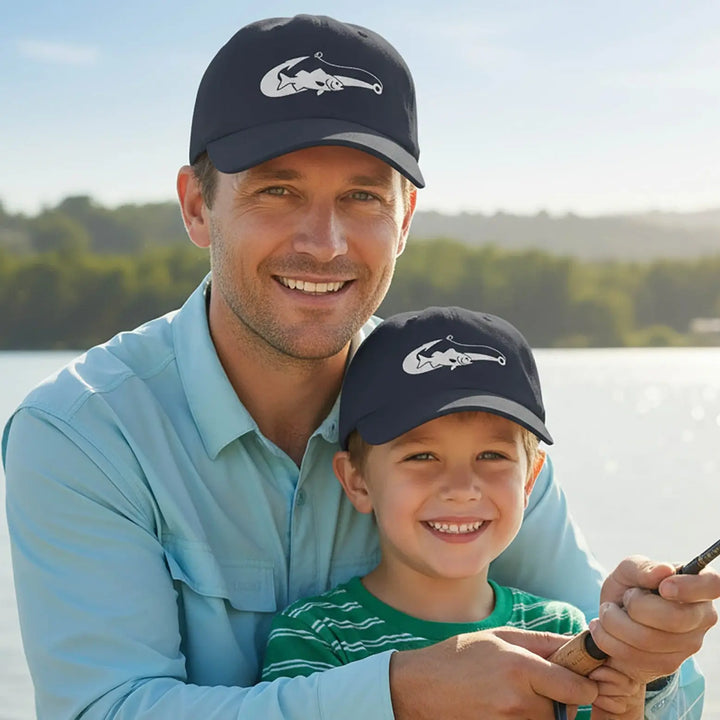 Man and child wearing matching navy hats with a logo, holding a fishing rod by a body of water.